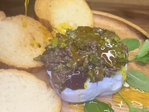 Chopped olive paste drizzled with golden oil atop white cheese, surrounded by bread slices and fresh herbs